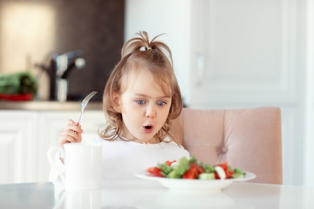 Menina criança de expressão emocional comendo salada de vitamina de vegetais crus frescos na cozinha branca. conceito de comida saudável para crianças