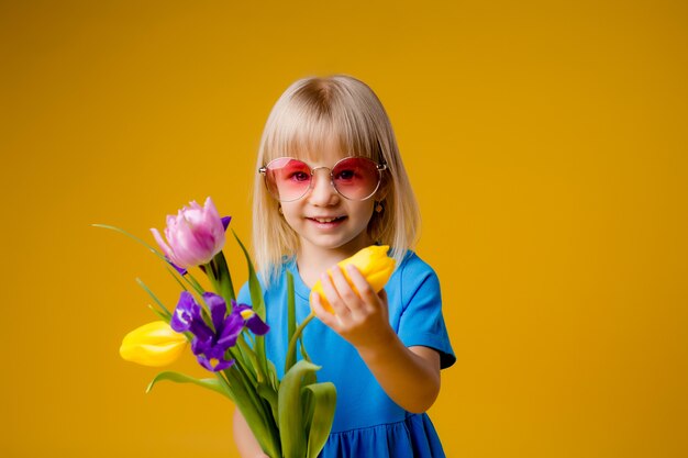 Foto menina criança em óculos de sol em roupas azuis, sorrindo e segurando um buquê de flores sobre fundo amarelo de isolar