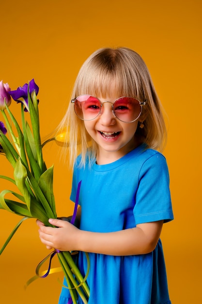 Foto menina criança em óculos de sol em roupas azuis, sorrindo e segurando um buquê de flores sobre fundo amarelo de isolar