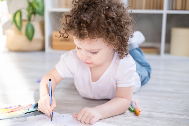 Menina criança encaracolada desenhando com marcadores coloridos no chão na sala de estar em casa