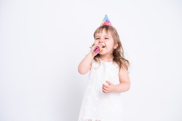 Menina criança engraçada no vestido branco e chapéu de aniversário soprando apito na superfície branca.