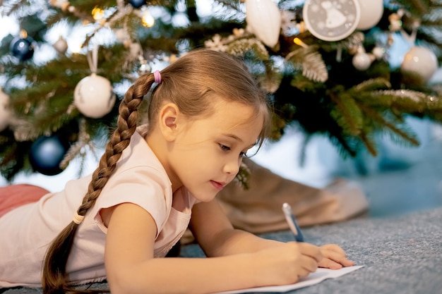 Menina criança escreve carta Papai Noel e sonha com uma árvore de ano novo de fundo de presente dentro de casa. Feliz Natal e Boas Festas.