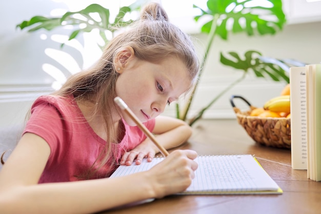 Menina criança estudando em casa, sentada à mesa, escrevendo no caderno da escola. lição de casa, ensino à distância, escola moderna e educação