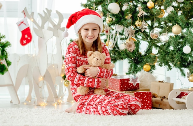 Menina criança feliz abraçando o ursinho de pelúcia brinquedo em casa com a árvore de Natal no fundo. Garoto fofo com presentes no ano novo