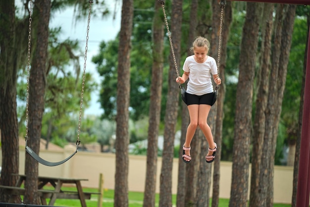 Menina criança feliz brincando sozinha voando alto em balanços no dia ensolarado de fim de semana de verão Segurança e recreação no conceito de playground