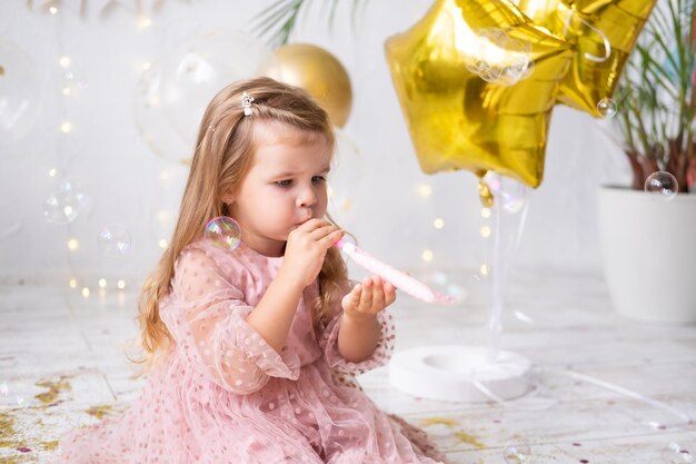 Menina criança feliz com cabelo comprido em um vestido rosa, soprando um apito e comemorando seu aniversário