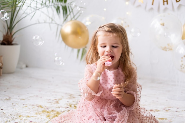 Menina criança feliz com cabelo comprido em um vestido rosa, soprando um apito e comemorando seu aniversário