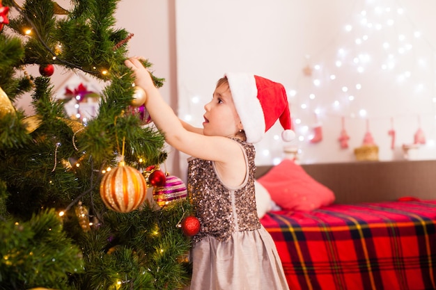 Menina criança feliz decorando a árvore de Natal em casa. Menina usa vestido festivo e chapéu de Papai Noel