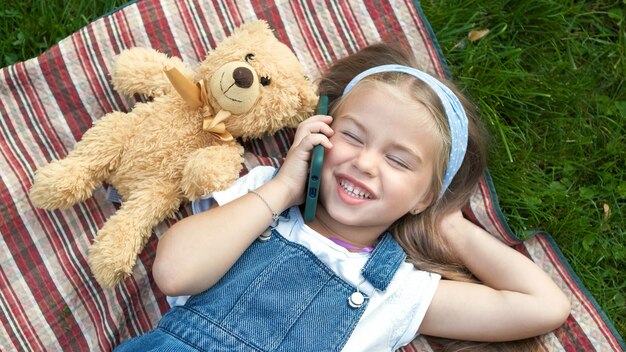 Menina criança feliz deitado sobre um cobertor no gramado verde no verão com seu ursinho de pelúcia falando no celular.