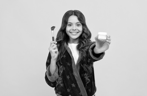 Menina criança feliz em roupão de banho aconchegante, preparando-se com pincel de pó apresentando produto