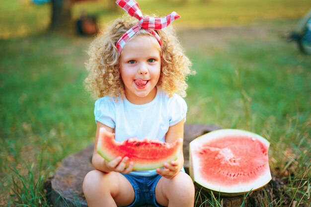 Menina criança feliz encaracolado sentado no tronco e comendo melancia ao ar livre no parque primavera contra fundo desfocado ensolarado natural