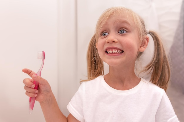 Menina criança feliz escovando os dentes no banheiro com uma escova de dentes. Se olha no espelho e sorri.