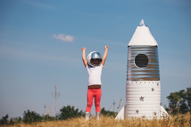 Menina criança feliz vestida com uma fantasia de astronauta, brincando com um foguete feito à mão. Verão ao ar livre.