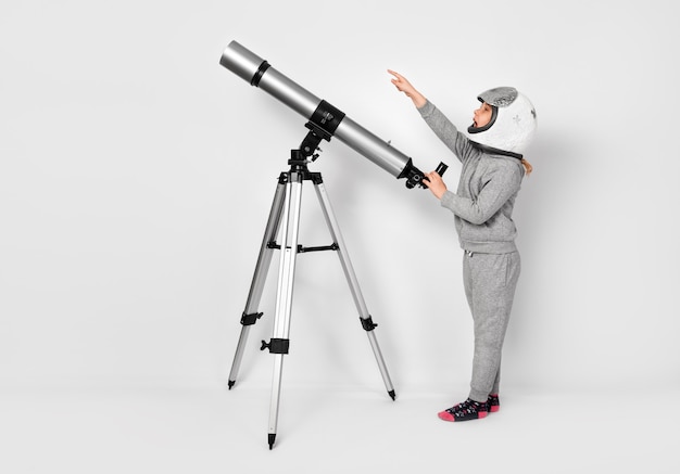 Menina criança feliz vestida com uma fantasia de astronauta em pé ao lado do telescópio.