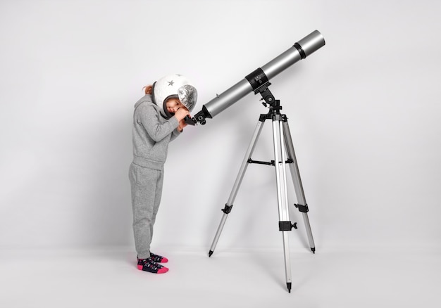 Menina criança feliz vestida com uma fantasia de astronauta olha através de um grande telescópio.