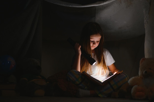 Foto menina criança lendo com livro e lanterna e ursinho de pelúcia na barraca antes de ir para a cama