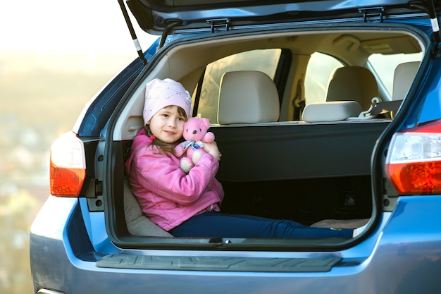 Menina criança muito feliz brincando com um ursinho de pelúcia de brinquedo rosa no porta-malas de um carro.