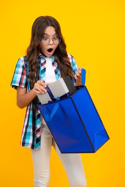 Menina criança segura sacola de compras desfrutando de venda isolada em fundo amarelo Retrato de menina adolescente está pronto para ir às compras Menina adolescente surpresa chocada