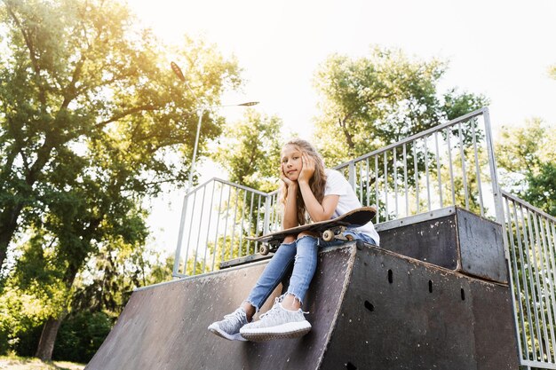 Menina criança sentada com prancha de skate na rampa esportiva Equipamentos esportivos para crianças Adolescente ativo com prancha de skate no parque de skate