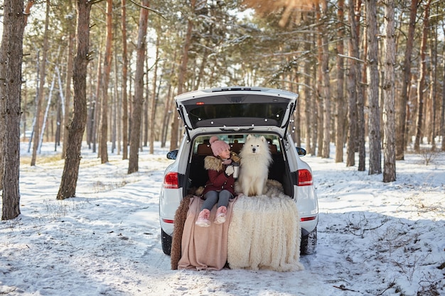 Menina criança sentada no porta-malas do carro com seu animal de estimação, um cachorro branco samoieda, no inverno em uma floresta de pinheiros nevados, uma menina bebendo chá em uma garrafa térmica