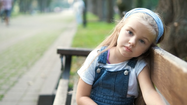 Menina criança séria sentada sozinha num banco no parque de verão.