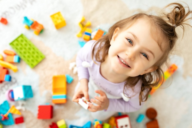 Menina criança sorridente sentada no chão da sala das crianças e brincando com blocos coloridos
