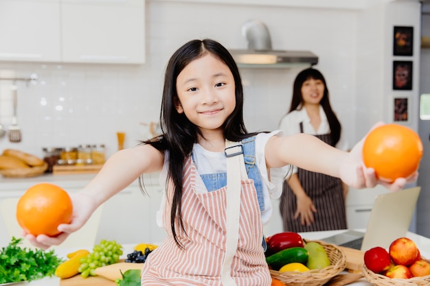 Menina criança sorrindo com a mão dando ou mostra fruta laranja na cozinha de casa com a mãe