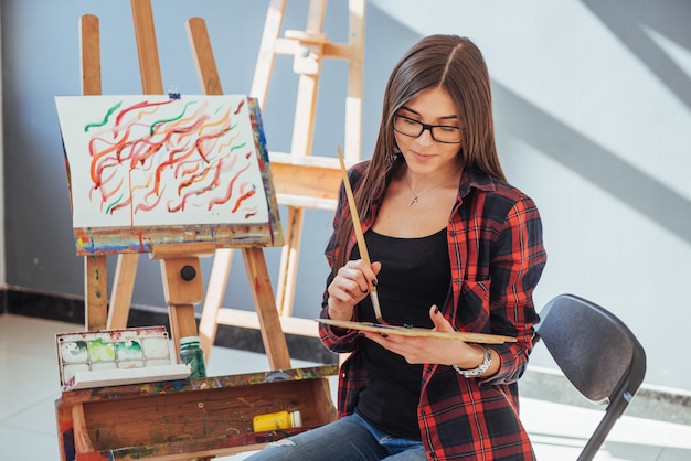 Foto menina criativa pintor pensativo pinta uma imagem colorida na lona com cores de óleo na oficina.