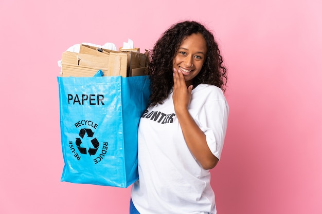 Menina cubana adolescente segurando um saco de reciclagem cheio de papel para reciclar isolado na parede rosa olhando para cima enquanto sorri