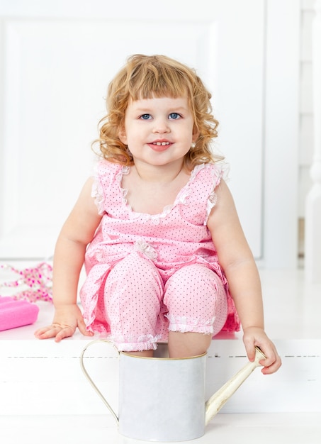 Menina curly bonito pequena em um vestido cor-de-rosa com bolinhas que sorri, sentando-se no estilo branco de Provence do patamar