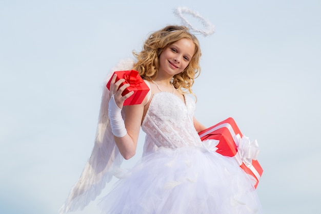 Menina da criança usando vestido branco de fantasia de anjo e asas de penas. Anjinho Cupido fofo com um presente