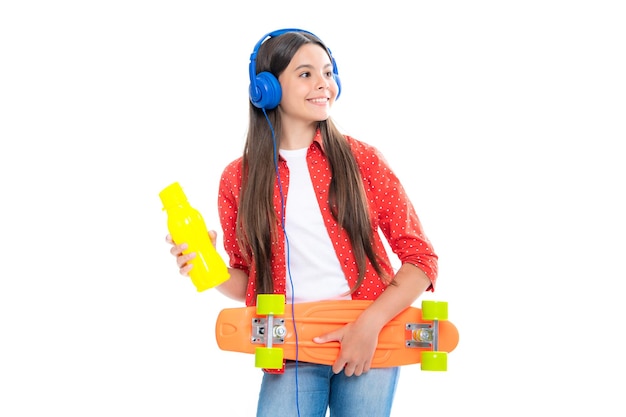 Menina da escola adolescente com garrafa de água de skate e fones de ouvido no fundo isolado do estúdio Estilo de adolescente urbano de tendência de primavera de estudante Retrato de menina adolescente sorridente feliz