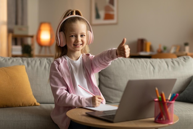 Menina da escola mostrando os polegares para o laptop usando fone de ouvido interno