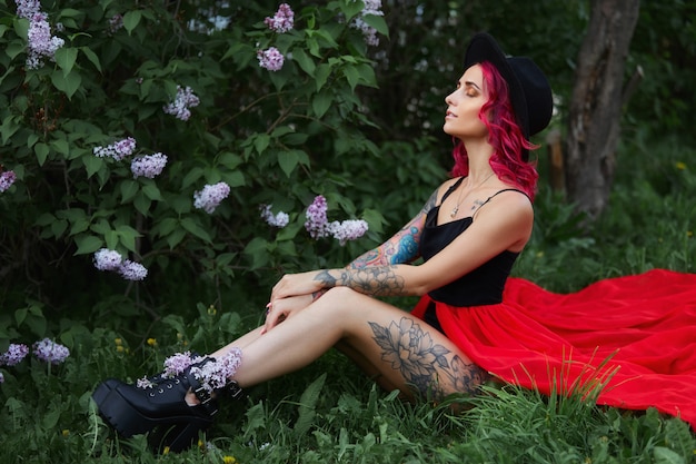 Menina da moda com cabelo ruivo e vocação de chapéu grande, retrato de primavera em cores lilás no verão. Lindo vestido rosa vermelho, tatuagens no corpo de uma mulher. Maquiagem brilhante, coloração de cabelo profissional