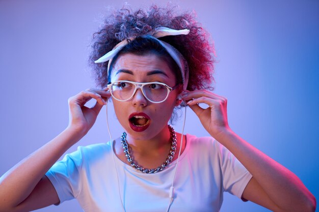 Menina da moda com cachos afro, rosto chocado e boca aberta usando fones de ouvido na parede de neon