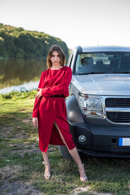 Menina da moda no elegante vestido vermelho em pé perto de seu carro moderno e relaxe a natureza no dia de verão