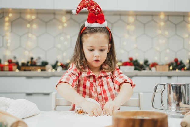 Menina de 3 anos com boné de Natal vermelho e camisa quadriculada preparando massa para biscoitos de gengibre na cozinha