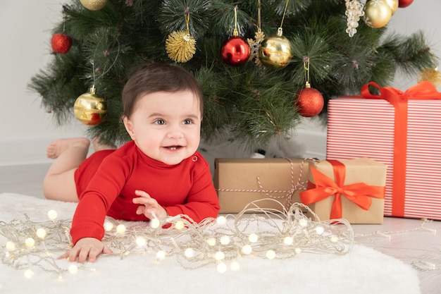 Menina de 6 meses em body vermelha sob a árvore de Natal com presentes de caixas de presente.