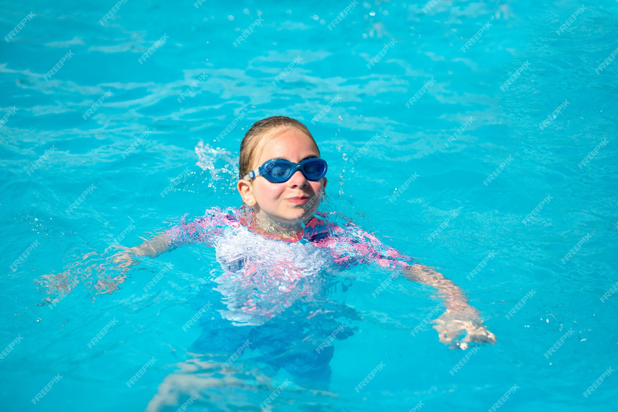 Menina de 8 anos, em um maiô brilhante e óculos azuis, nadando em uma  piscina sob o sol com água azul, feliz e feliz