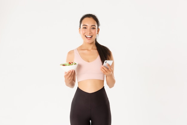 Menina de aptidão asiática magro sorridente em roupas esportivas, segurando salada e telefone celular, usando o aplicativo de lembrete de comer, aplicativo de controle de dieta, verificação de calorias.