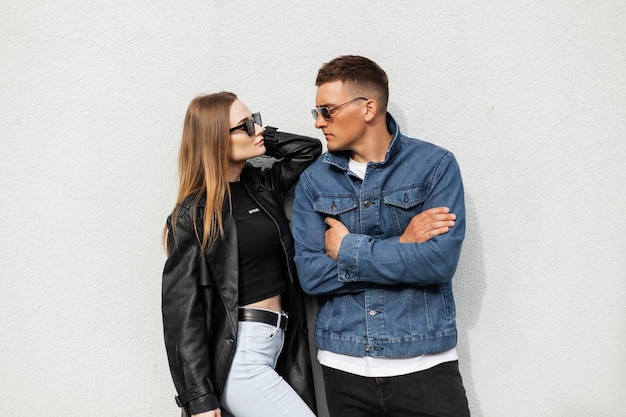 Menina de beleza elegante e um homem bonito de moda com óculos de sol legais em roupas de rua casuais da moda ficam perto de uma parede branca na rua Casal urbano de moda em roupa jeans elegante