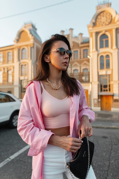 Menina de beleza hipster modelo elegante com óculos de sol em uma camisa rosa na moda e zíperes superiores em sua bolsa de moda e andando na cidade