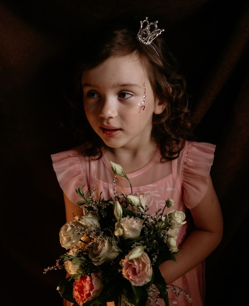 Menina de cabelo encaracolado com uma fita na cabeça com uma coroa e um vestido festivo segurando um buquê de rosas em um fundo marrom