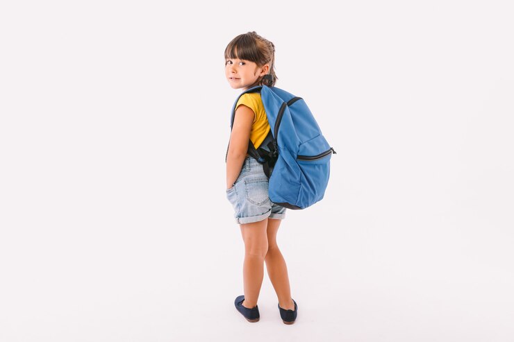Menina de cabelo preto vestida com macacão jeans e camiseta azul, com mochila pronta para voltar às aulas, de costas para a escola, sobre fundo branco