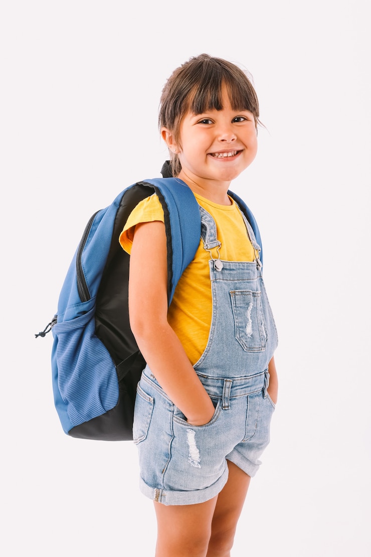 Foto menina de cabelo preto vestida com um macacão jeans e uma camiseta azul, com uma mochila pronta para voltar às aulas, ao seu lado, sobre fundo branco