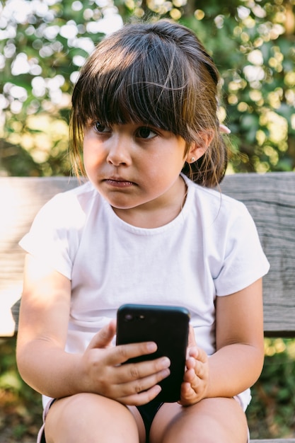 Menina de cabelo preto, vestindo uma camiseta branca, sentada num banco de um parque, olhando para o celular, com um gesto sério.