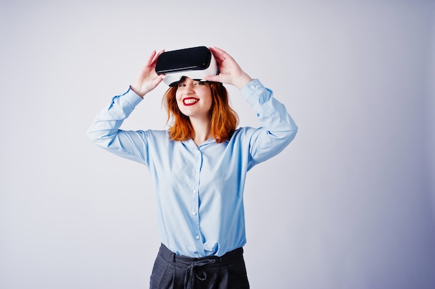 Menina de cabelo vermelha na blusa azul com os vidros do vr isolados no branco.