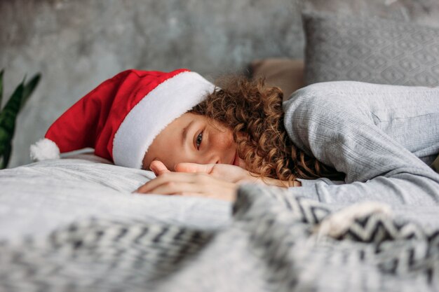 Menina de cabelos cacheados tween linda no chapéu de Papai Noel e pijama, sentada na cama com travesseiro, tempo de manhã de Natal