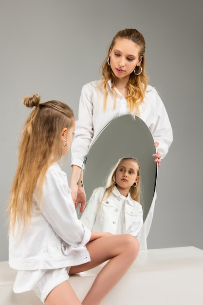 Menina de cabelos compridos em roupas brancas olhando seriamente para ela refletindo enquanto a irmã mais velha carregava um espelho