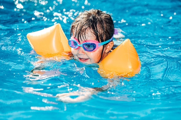 Menina de cabelos pretos sorrindo e nadando na piscina com mangas e óculos de natação